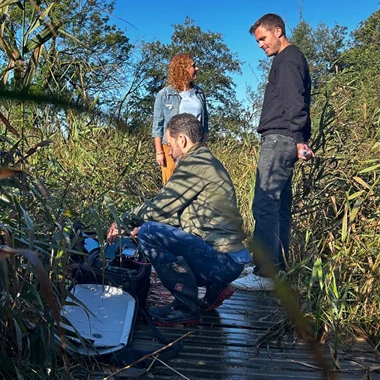 Drie mensen tijdens een fotoshoot in een natuurgebied, staand en knielend op een houten vlonder tussen het riet.