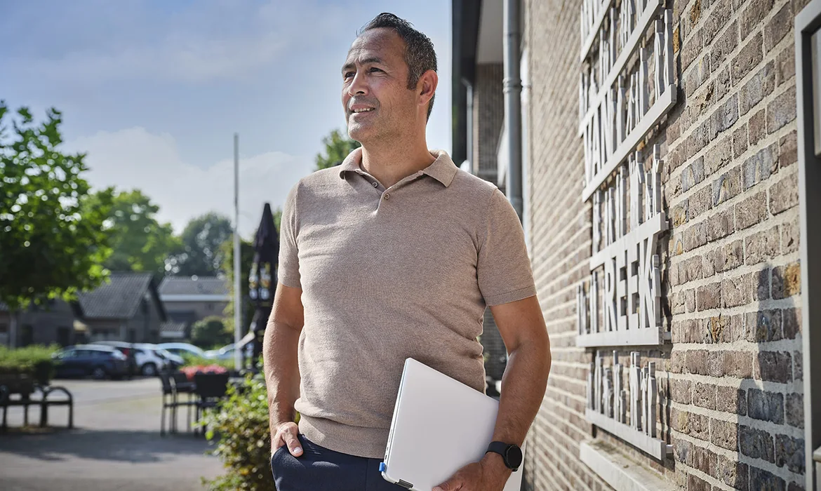 Een man met een laptop onder zijn arm kijkt vooruit terwijl hij ontspannen bij bakstenen muur staat in een sfeervolle dorpsomgeving.