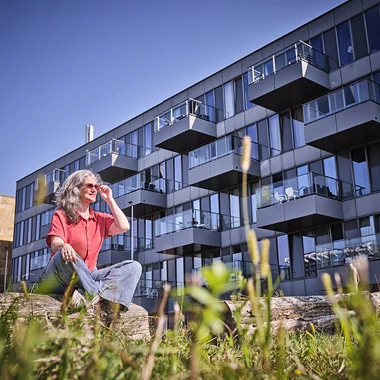 Vrouw zit ontspannen in het groen voor een modern appartementencomplex met glazen balkons.