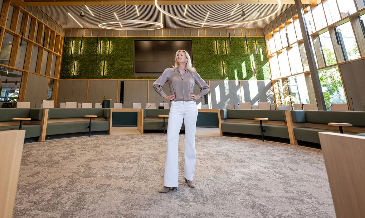 Een vrouw staat zelfverzekerd in een moderne, duurzame vergaderruimte met groene wandpanelen en houten accenten.