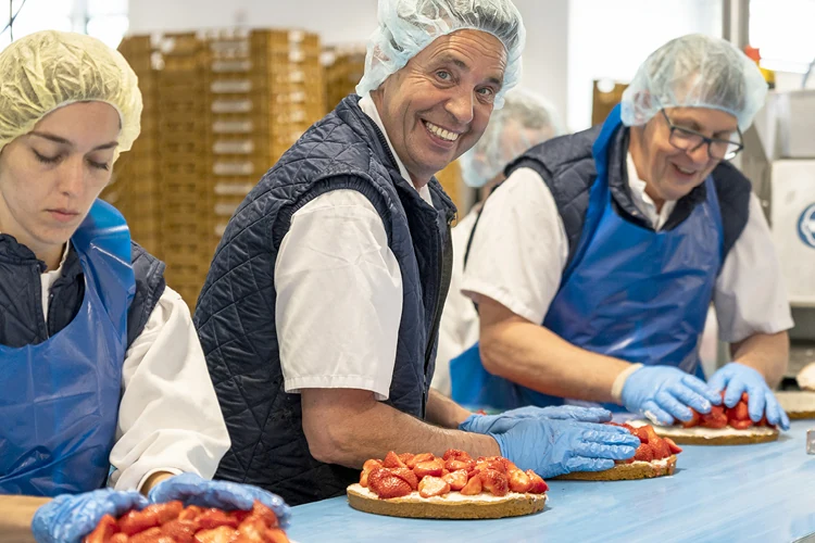 Medewerkers in een bakkerij bereiden aardbeientaarten voor, met haarnetjes, handschoenen en blauwe schorten; een man in het midden lacht naar de camera.