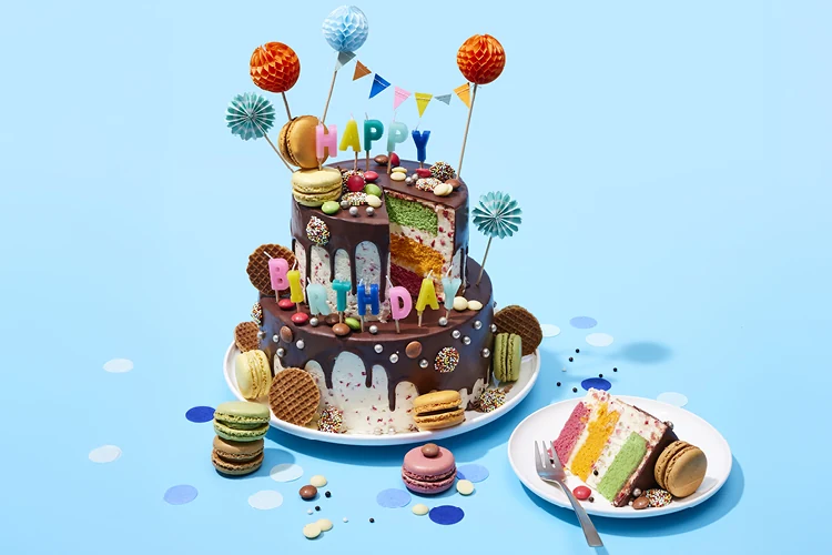 Twee verdiepingen tellende feestelijke verjaardagstaart met chocoladeglazuur, macarons, stroopwafels en kleurrijke decoraties, met een uitgesneden stuk op een bord ernaast.