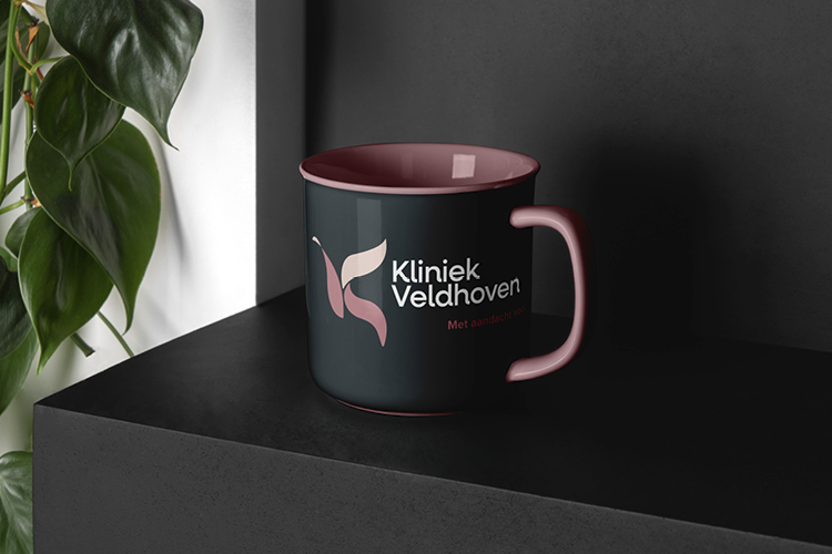 Donkere mok met roze binnenkant en handvat, bedrukt met het logo van Kliniek Veldhoven en de slogan