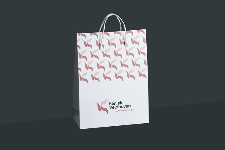 Witte papieren tas met het logo en patroon van Kliniek Veldhoven, voorzien van het motto