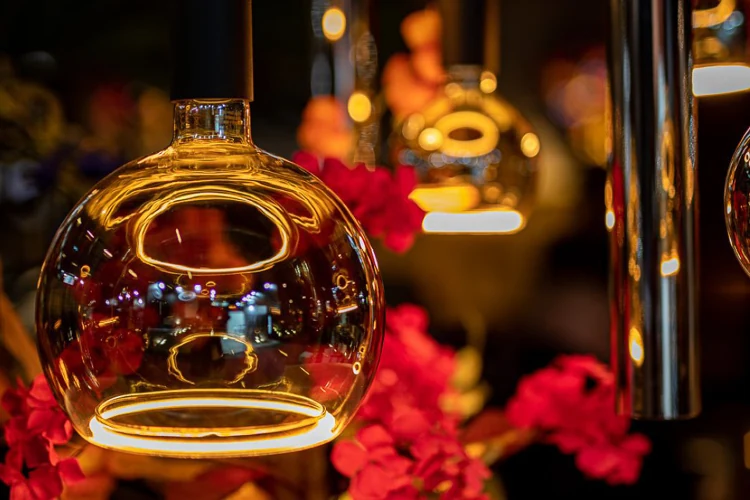 Close-up van een glazen designlamp met warme verlichting, omringd door rode bloemen in een sfeervolle setting.
