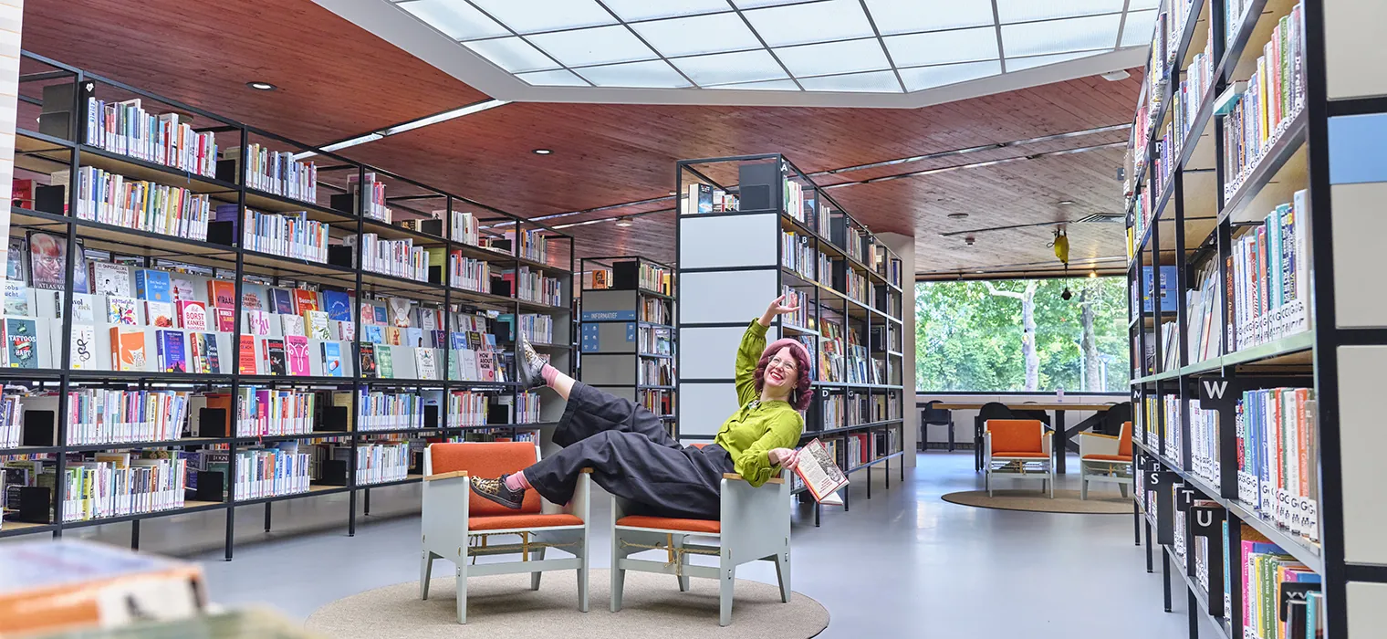 Vrolijke persoon met rode hoed en groene blouse ligt speels achterover in een stoel in een moderne bibliotheek, omringd door boekenrekken en grote ramen met uitzicht op groen.